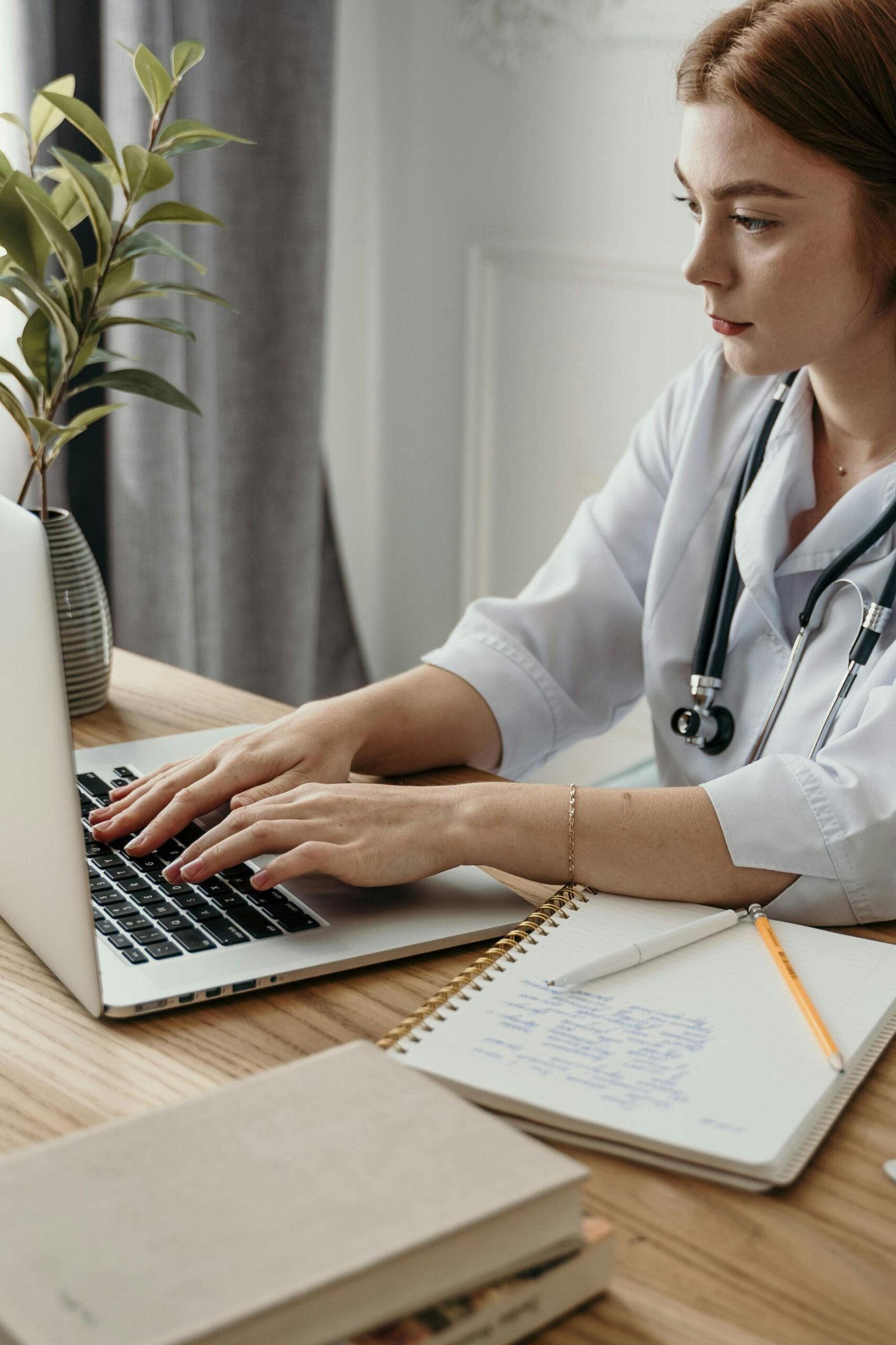 A professional female doctor typing on a laptop in her office with medical notes and stethoscope.